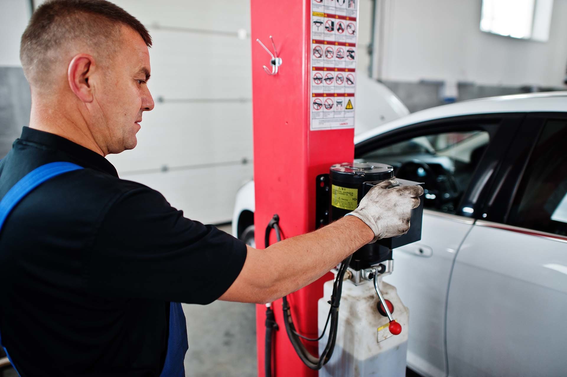 Ein Werkstattmitarbeiter bedient eine rote Hebebühne zur Wartung eines weißen Autos – AH Werkstatt Service aus Neuhofen bietet spezialisierte Dienstleistungen für Hebebühnen und Werkstattausrüstung.