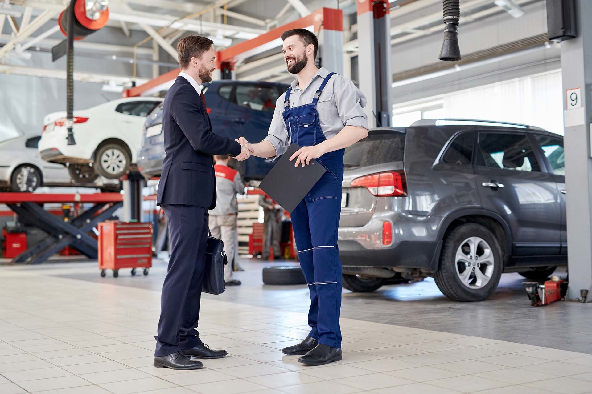 Zwei Männer schütteln sich in einer Autowerkstatt die Hände: ein Geschäftsmann in Anzug und ein Mechaniker in Overalls, mit Fahrzeugen und Werkstattausrüstung im Hintergrund, symbolisch für die Serviceleistungen von AH Werkstatt Service für Hebebühnen und Werkstattausrüstung.