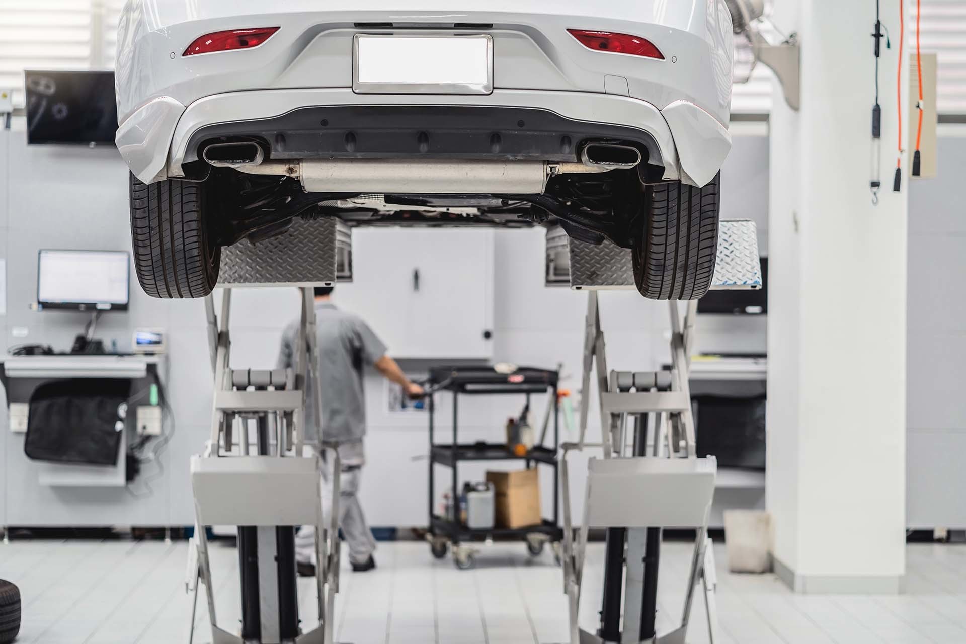 Weißes Auto auf einer Hebebühne in einer modernen Werkstatt, mit einem Mechaniker im Hintergrund – zeigt die Hebetechnik und Werkstattausrüstung, die AH Werkstatt Service anbietet.