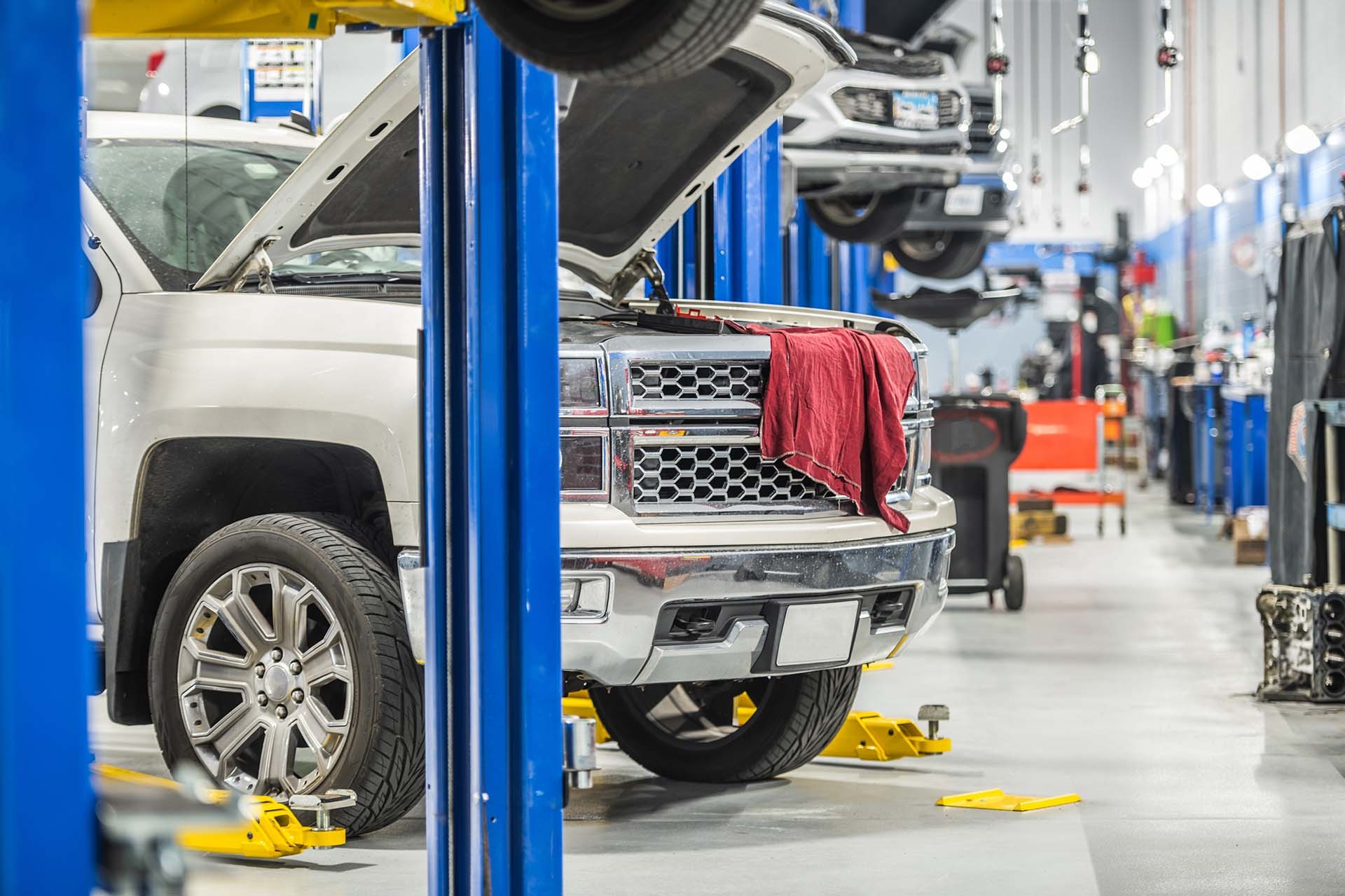 In einer Werkstatt steht ein weißer Lkw auf einer blauen Hebebühne mit geöffneter Motorhaube, umgeben von Werkstattausrüstung – ein Beispiel für die Hubtechnik und Serviceleistungen von AH Werkstatt Service, spezialisiert auf Hebebühnen und Werkstattausrüstung.
