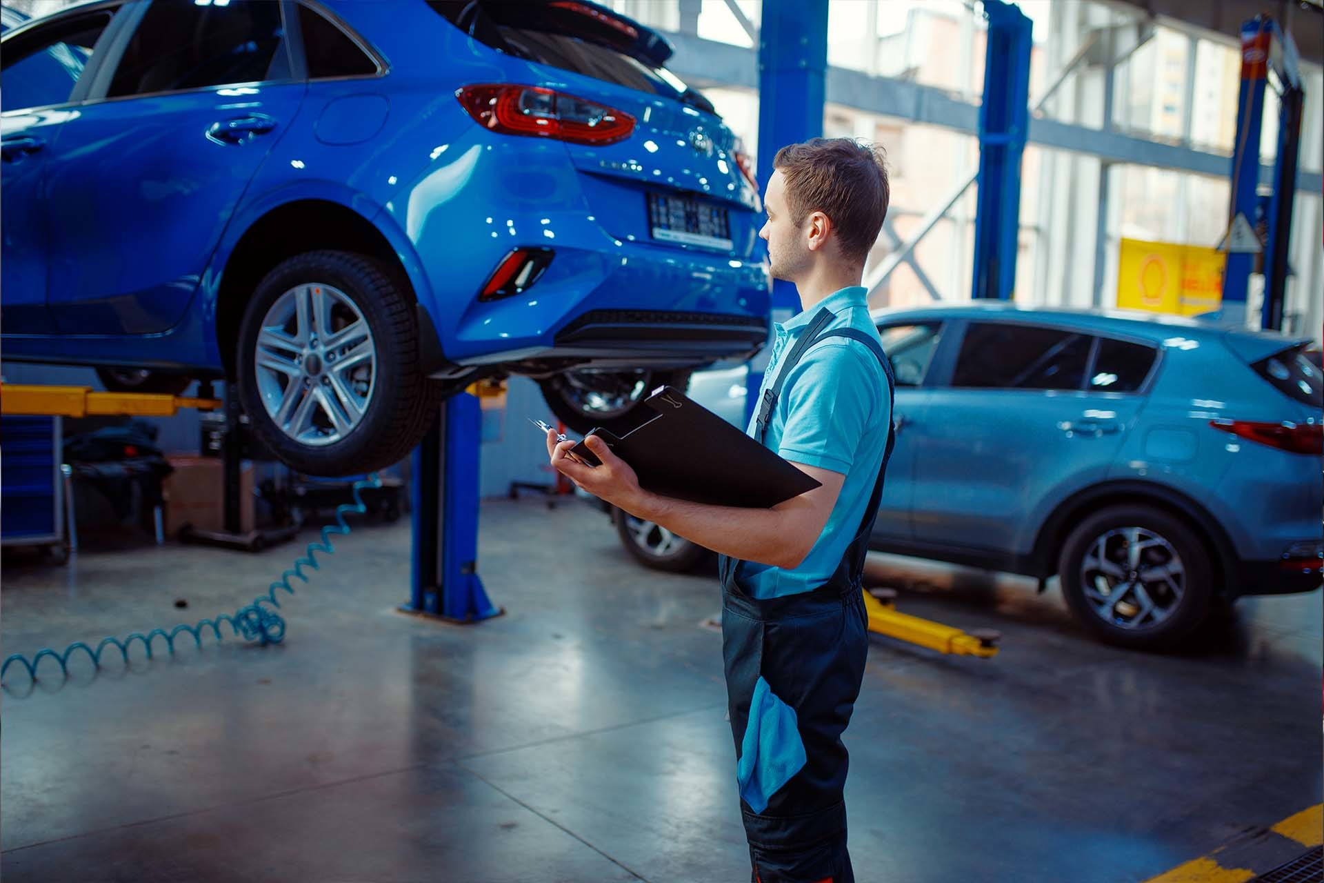 Mechaniker in einer Werkstatt, der ein blaues Fahrzeug auf einer Hebebühne kontrolliert, mit einem weiteren Auto im Hintergrund – repräsentiert die Dienstleistungen von AH Werkstatt Service für sichere und zuverlässige Hubtechnik in Werkstätten.